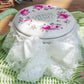 Vintage white tin container with pink floral design and 'Country Charm' text on green wicker basket, adorned with white lace