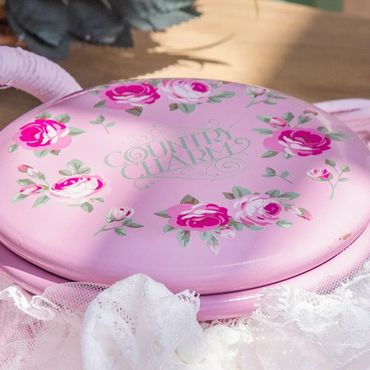 Pink round tin with floral design and 'Country Charm' text on wooden surface with white lace fabric