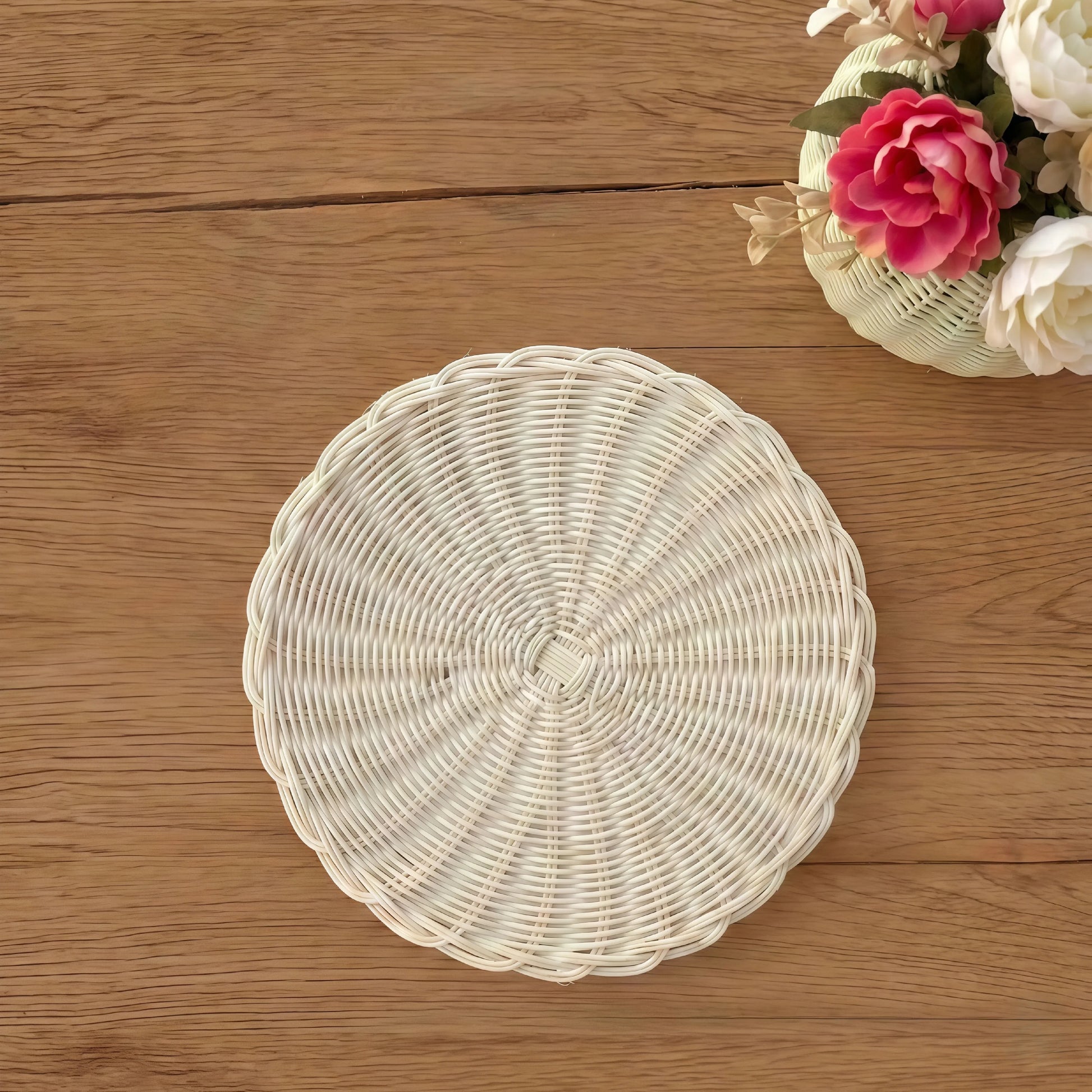 White round woven wicker placemat centered on a brown wood surface next to a vase with pink and cream flowers.