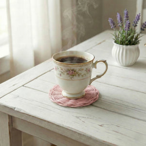 Steaming cup of coffee on a pink coaster with a small potted plant in the background.