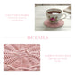 Teacup with floral design on a pink woven coaster on a wooden table, with text about the product.