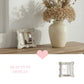 Antique white distressed mirror on a wood shelf next to a cream vase with dried flowers and a pink heart icon.