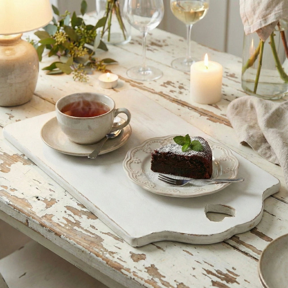 Antique white rectangular serving board with a heart-shaped handle, styled on a rustic wooden table with a steaming cup of tea, a slice of chocolate cake on a decorative plate, and lit candles.