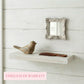 Antique white mirror on a plank wall above a floating shelf holding a wooden bird figurine and a brown comb.