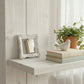 Antique white mirror leaning on a whitewashed shelf with books, a bird figurine, and a potted plant.