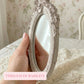 Hand holding a small antique white oval mirror with a distressed ornate frame against a floral wallpaper background.