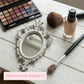 Ornate white oval mirror, makeup palette, foundation bottle, and brushes on a rustic white wooden surface.