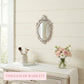 Ornate oval mirror on a shiplap wall above a vanity with makeup brushes, a glass tray, and pink flowers in a vase.