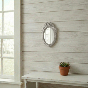 Ornate white oval mirror with a ribbon crest on a whitewashed wood wall above a small succulent and white table.