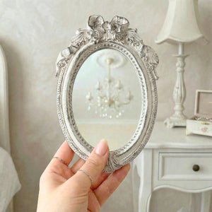 A hand holding an ornate antique white oval mirror with a ribbon crest, reflecting a chandelier in a bright room.