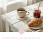 Antique white woven round coaster under a ceramic tea mug with a croissant and jam jar on a distressed white wood table by a window.