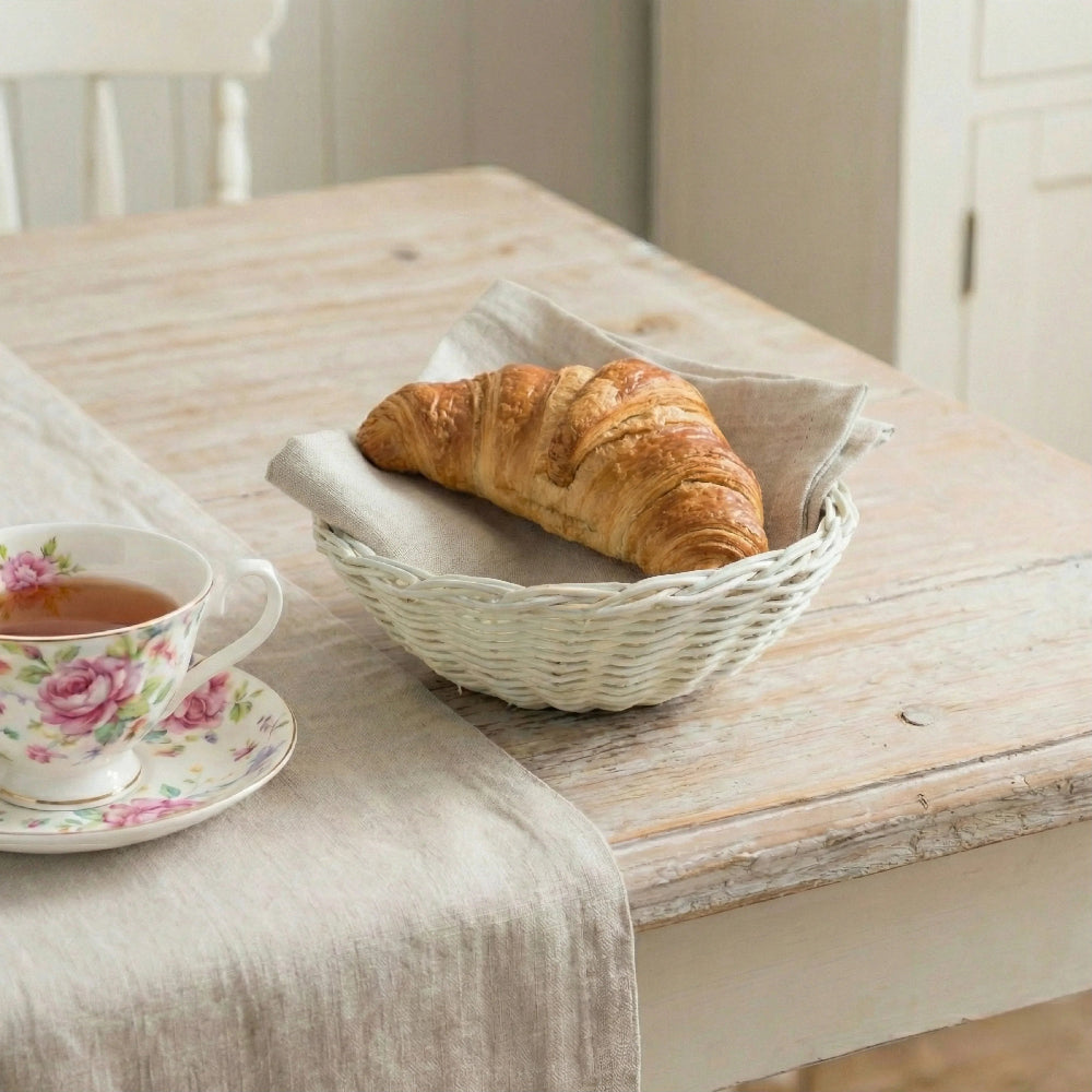 Antique white wicker basket holding a croissant on a linen napkin next to a floral teacup on a wooden table.