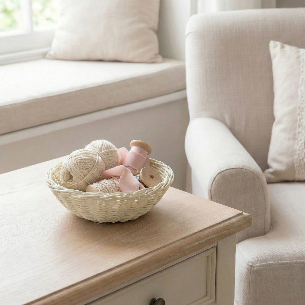 Antique white wicker basket filled with yarn and pink ribbon on a wooden table next to a beige armchair.