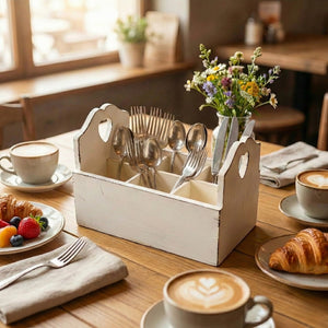 Antique white wooden cutlery caddy with six compartments and heart-shaped side handles, styled on a wooden cafe table with silverware, coffee, and croissants.