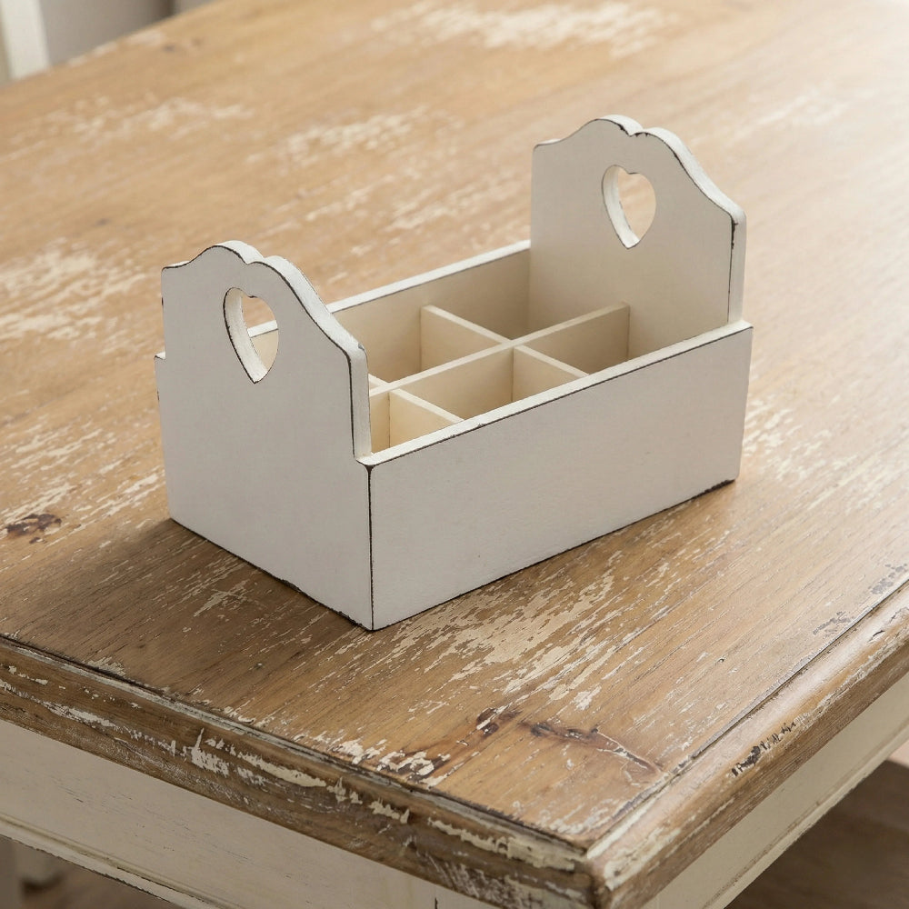 Antique white wooden flatware caddy with six compartments and heart-shaped handle cutouts, displayed on a rustic whitewashed wooden table.