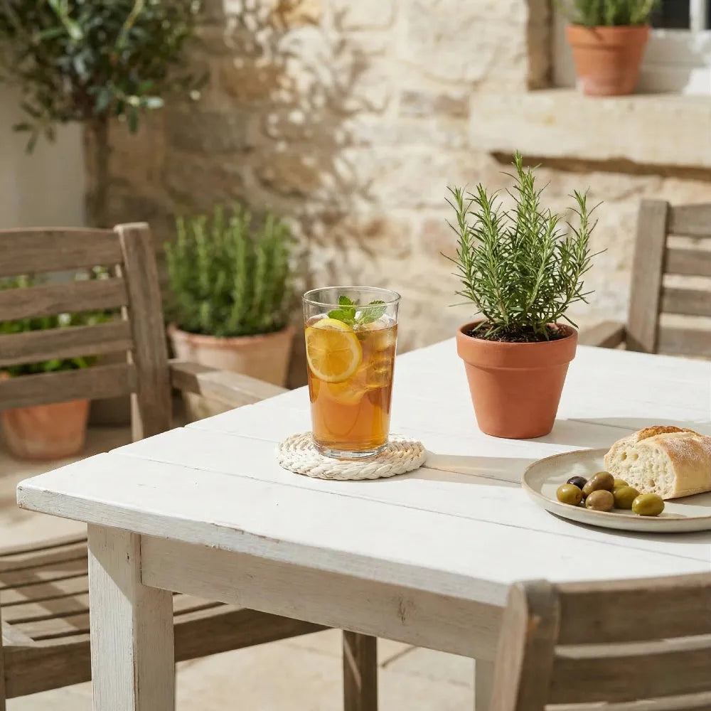 Outdoor setting with a table featuring a glass of iced tea, lemon slices, and a plate of bread and olives, with potted plants in the background.