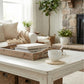 Antique white woven round coaster under a white mug on a distressed white coffee table in a bright, farmhouse-style living room with a fireplace and greenery.