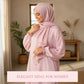 Woman wearing a pink hijab and polka dot Isdal Prayer dress in a home setting with plants and a wooden shelf.