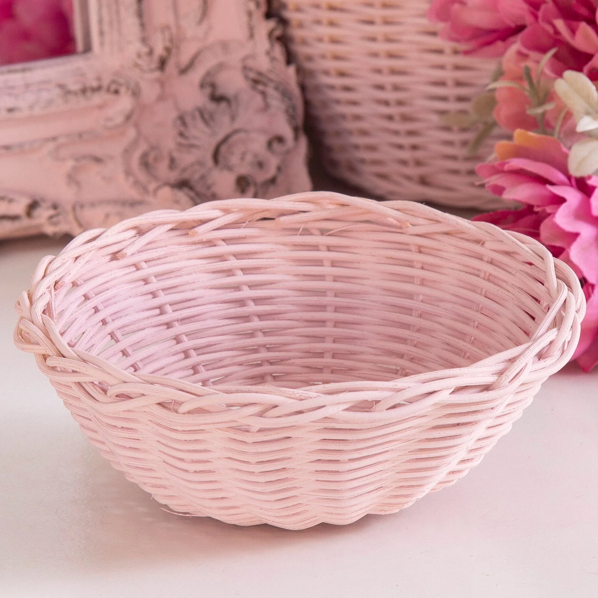 Pink wicker basket with braided rim, placed on a white surface near pink flowers and decorative frame