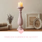 Decorative setup with a candle in a tall holder, vase with dried flowers, and framed pictures on a wooden surface.