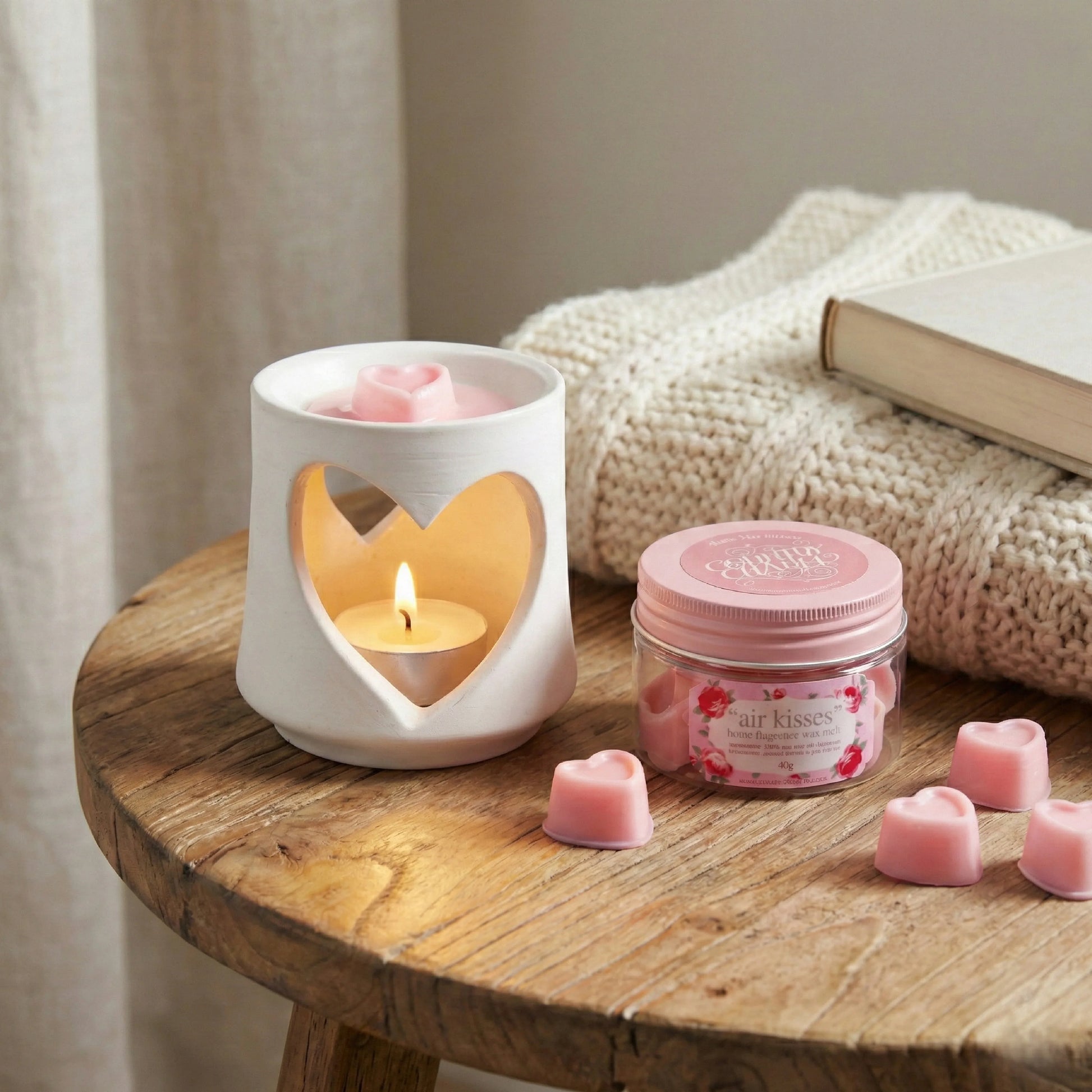 Candle warmer with a lit candle, jar of pink heart-shaped candles, and a book on a wooden surface.

