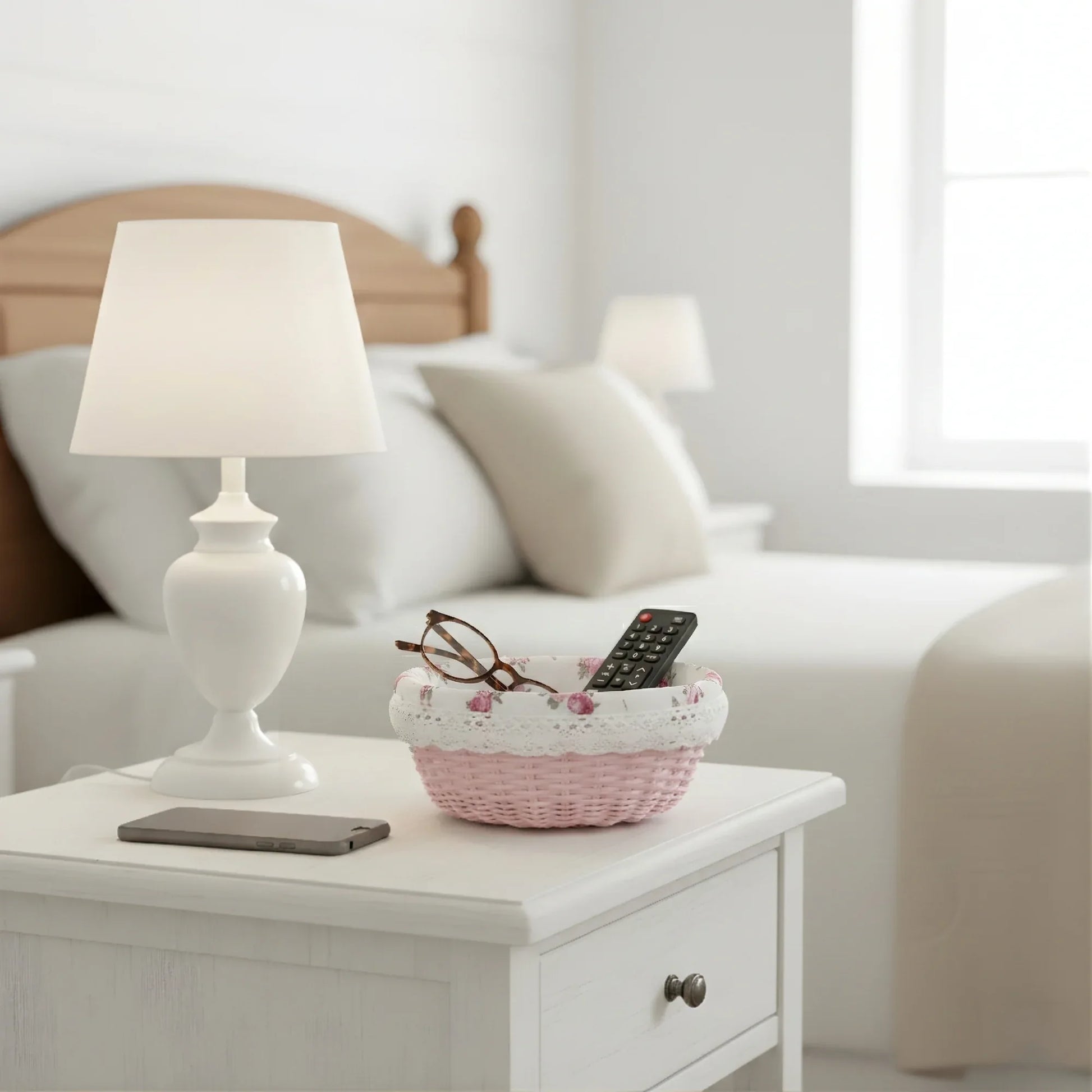 White lamp on a nightstand with a basket containing glasses and a remote control, in front of a bed.