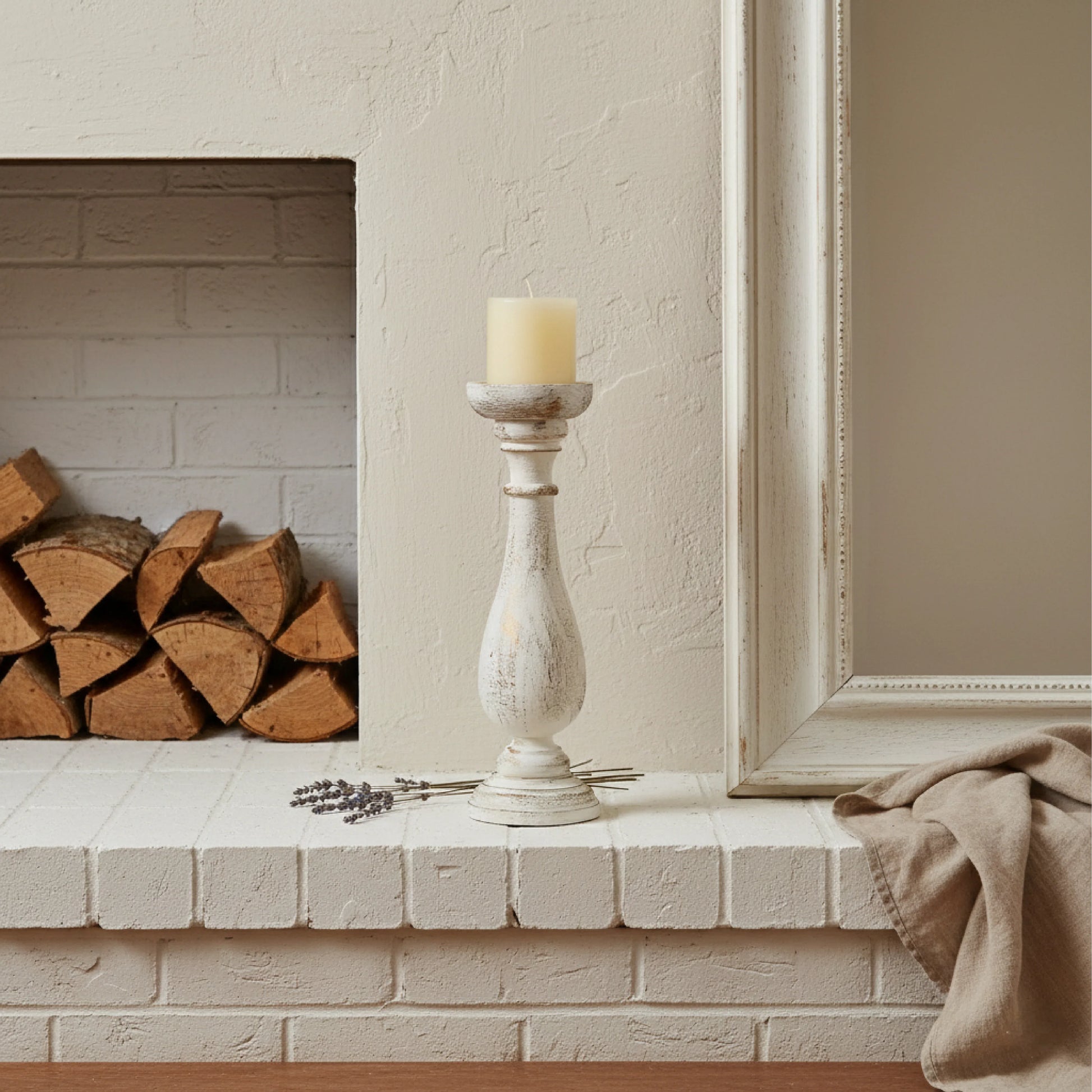 Candle in a decorative holder on a fireplace mantel with stacked logs and a framed picture.