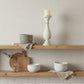Wooden shelves with ceramic bowls, a candle holder, and a cutting board against a neutral wall.