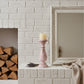 Pink candle holder with a candle on a white brick fireplace mantle, next to stacked firewood and a framed picture.