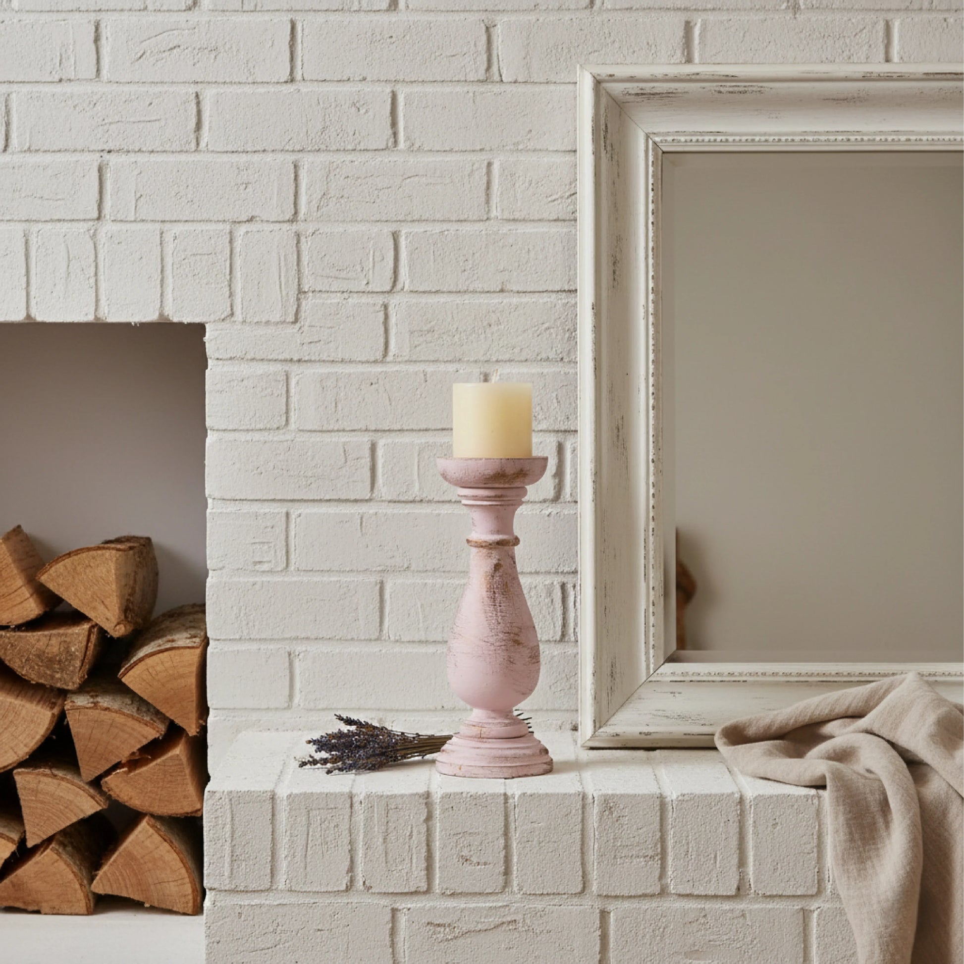 Pink candle holder with a candle on a white brick fireplace mantle, next to stacked firewood and a framed picture.