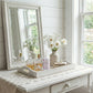 White dresser with mirror, flowers, and decorative items in a bright room.