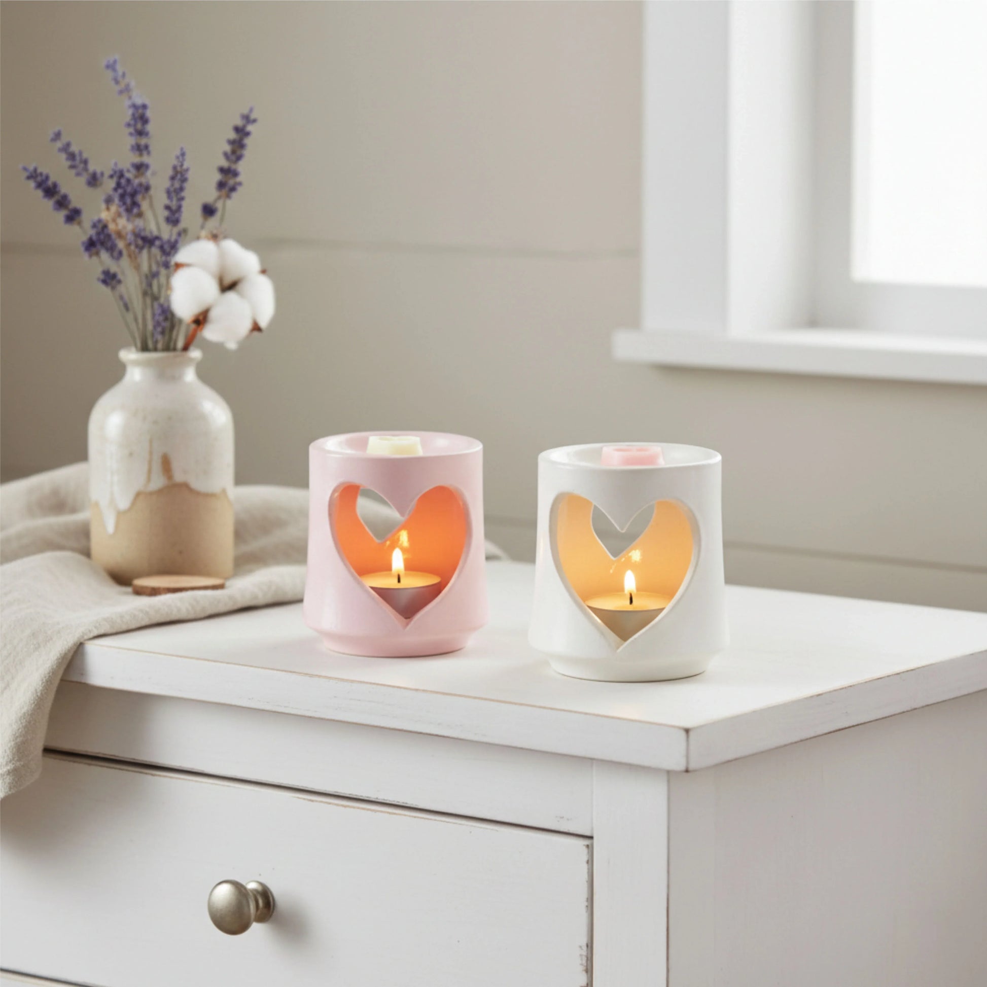 Two heart-shaped candle holders with lit candles on a white surface, accompanied by a vase with flowers.

