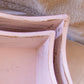 Close-up of two distressed light pink wooden trays with curved edges on a soft fabric background