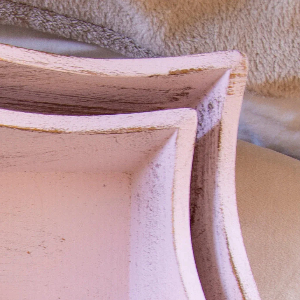 Close-up of two distressed light pink wooden trays with curved edges on a soft fabric background