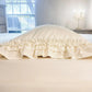 A cream-colored pillow with layered ruffled edges resting on a white mattress in front of a window.