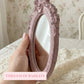 Hand holding a distressed pink ornate oval mirror over lace fabric with floral wallpaper background and pink text.