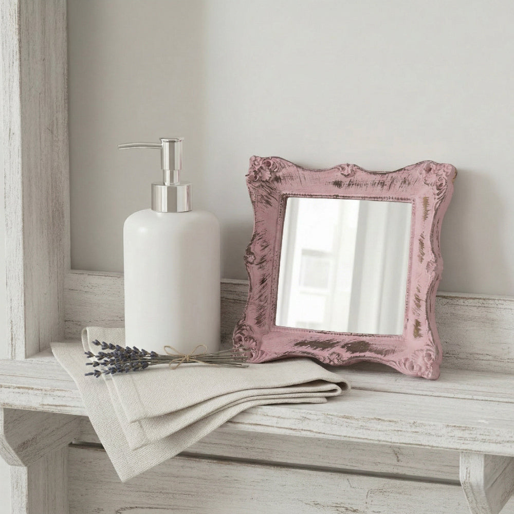Distressed pink ornate mirror on a white wooden shelf next to a soap dispenser and dried lavender on a linen cloth.