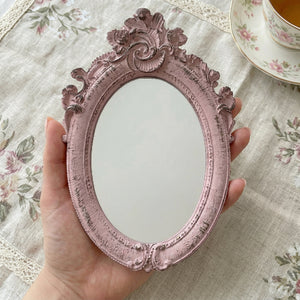 Hand holding a distressed pink ornate oval mirror over a floral tablecloth next to a teacup and saucer.