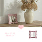Pink distressed mirror on a wood shelf beside a cream vase with dried flowers, accompanied by a pink heart icon.
