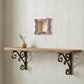 Small pink distressed mirror on a white wood wall above a rustic shelf with ornate scrolled iron brackets and a vase.