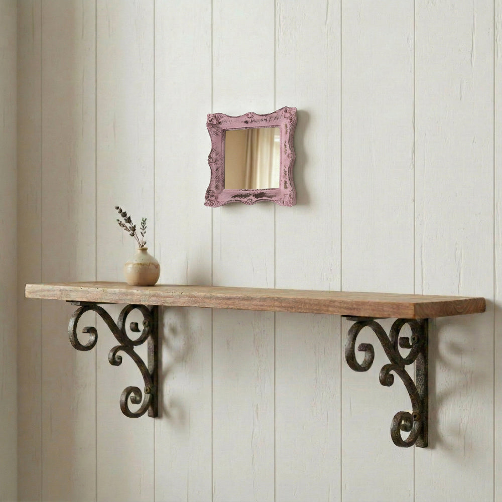Small pink distressed mirror on a white wood wall above a rustic shelf with ornate scrolled iron brackets and a vase.