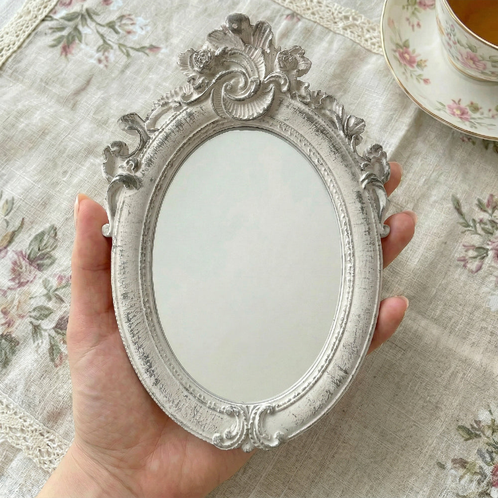 Hand holding a distressed white ornate oval mirror over a floral tablecloth next to a floral teacup.
