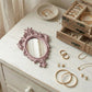 Ornate dusty pink mirror, gold jewelry, and a wooden jewelry box on a white distressed wooden dresser.
