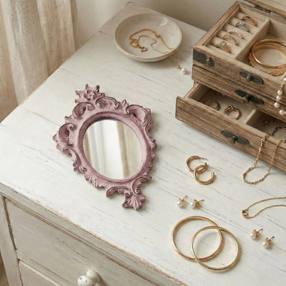 Ornate dusty pink mirror, gold jewelry, and a wooden jewelry box on a white distressed wooden dresser.