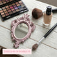 Ornate dusty pink mirror, eyeshadow palette, foundation, and makeup brushes on a white wooden surface.