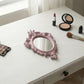 Ornate dusty pink mirror, lipstick, powder compact, and brushes on a white vanity desk.
