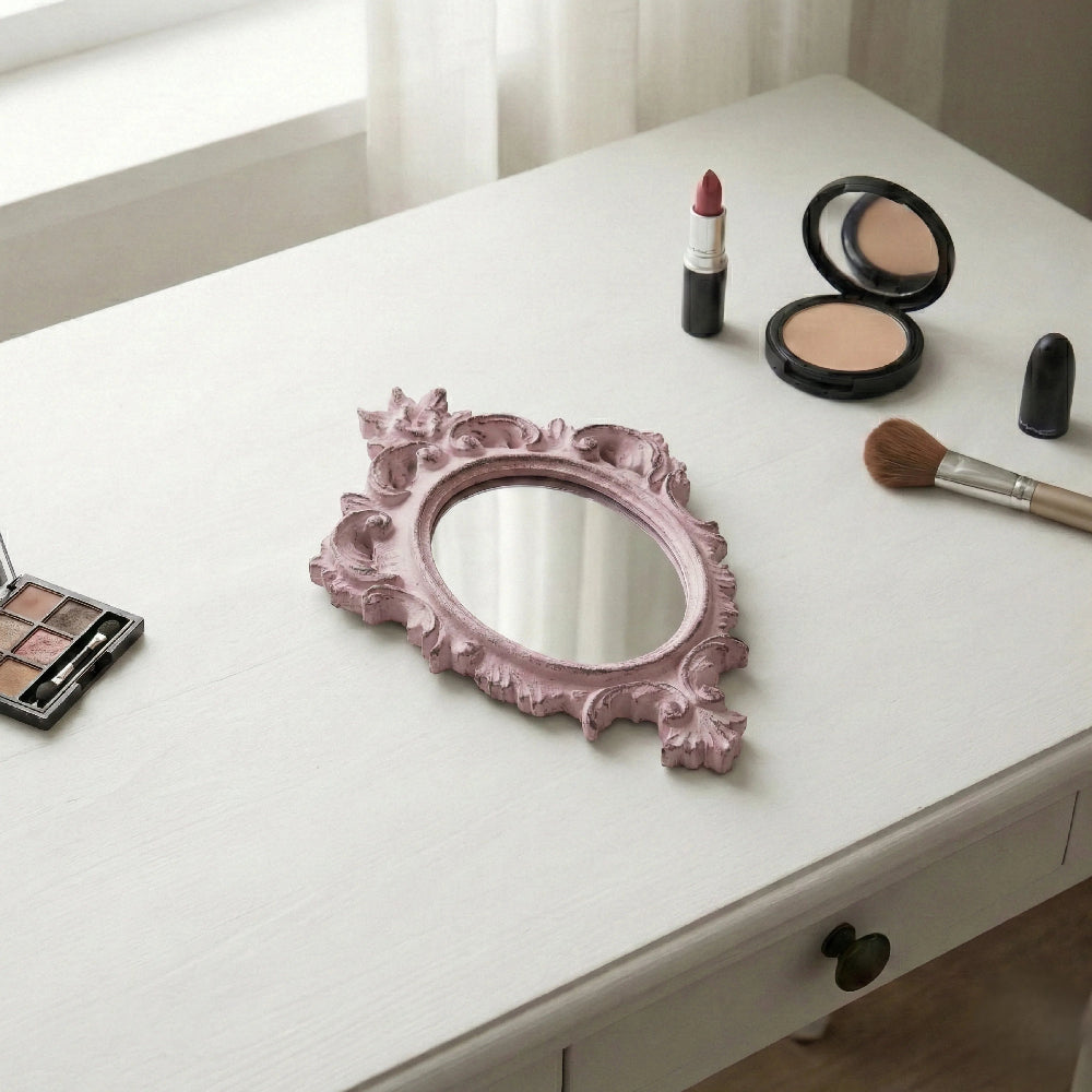 Ornate dusty pink mirror, lipstick, powder compact, and brushes on a white vanity desk.