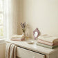 Stacks of folded linens, a lavender vase, and an ornate dusty pink mirror on a white dresser next to a lace curtain.