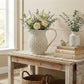 Decorative setting with a wooden table, wicker pitcher with flowers, books, and a vase.

