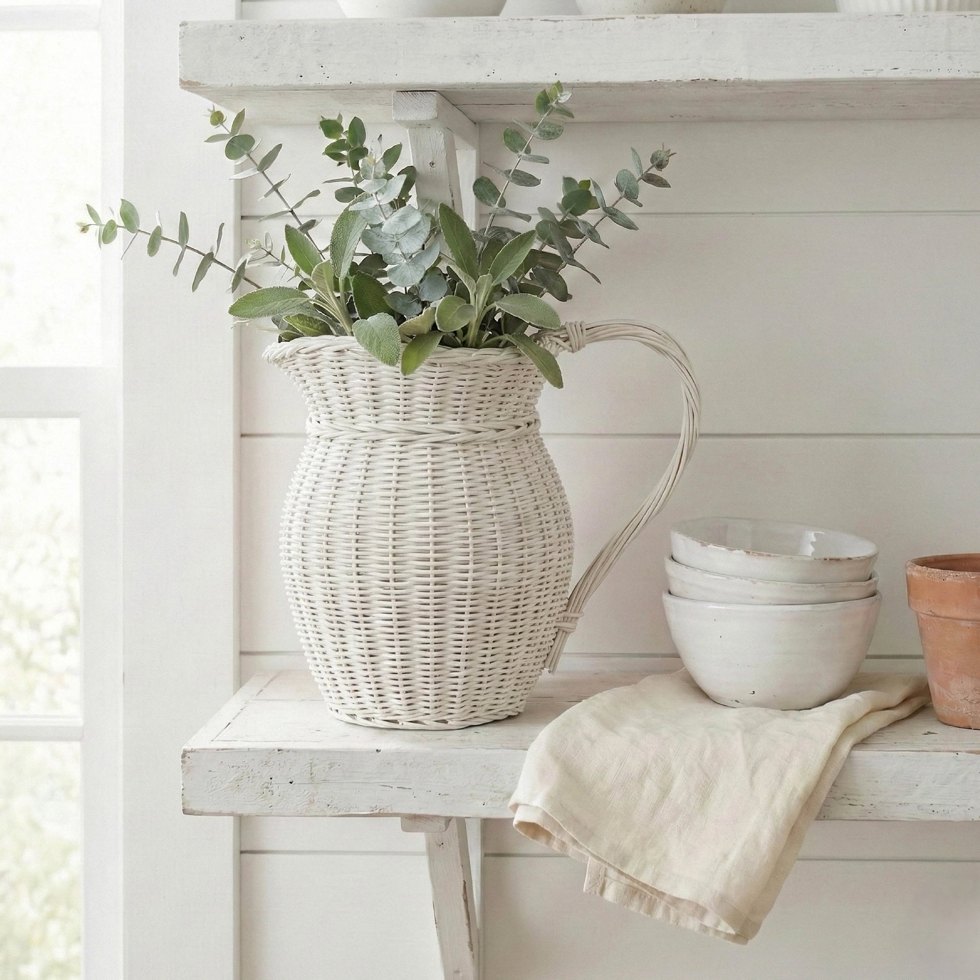 Wicker pitcher with greenery on a white wooden shelf with bowls and a towel.
